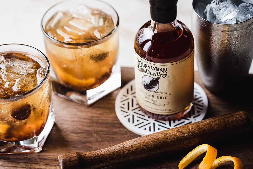 Bottle of Journeyman Distillery Rye whiskey next to two old-fashioned cocktails on a wooden serving tray